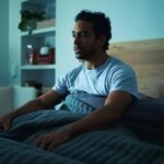 Insomnia, scared and man in bedroom at night for burnout, exhausted and fatigue for mental health. Tired, dark and person with fear, awake and afraid for sleepless, bad dream and nightmare in home