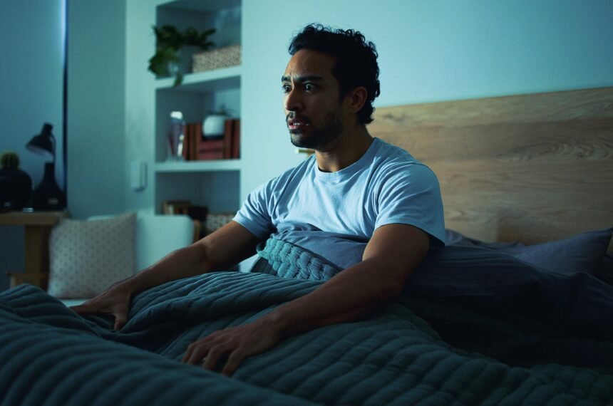 Insomnia, scared and man in bedroom at night for burnout, exhausted and fatigue for mental health. Tired, dark and person with fear, awake and afraid for sleepless, bad dream and nightmare in home